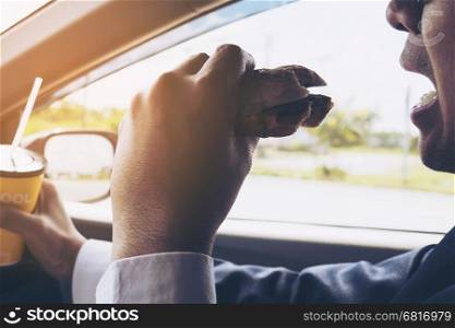 Man driving car while holding a cup of cold coffee and eating hamburger