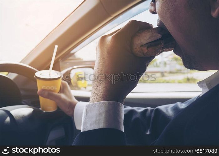 Man driving car while holding a cup of cold coffee and eating hamburger