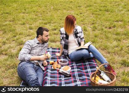 man eating banana looking her girlfriend reading book picnic