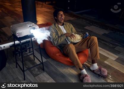 Man enjoy watching comedian film or show using video projector. Young laughing guy eating snack. Happy young man laughing during watch film