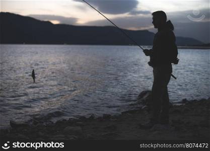 Man fishing on mountain lake. Sunrise low light. Casual clothes