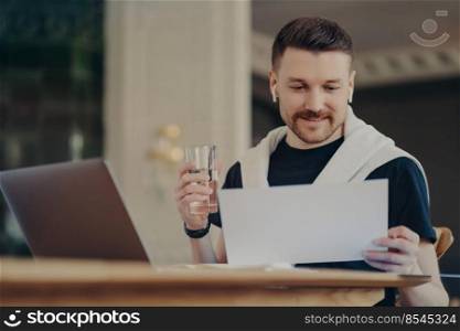 Man freelancer looking at document in front of laptop, sitting at home office at his work desk, holding glass of water and using earphones, young businessman in casual wear working remotely from home. Smiling attractive male freelancer sitting in front of laptop holding glass of water