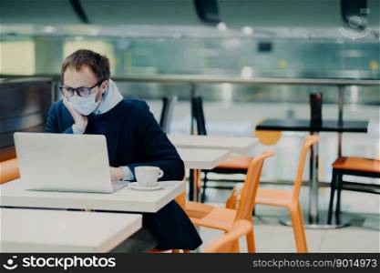 Man freelancer wears face mask to prevent spread of coronavirus and coughing, uses laptop computer for distant work, poses in cafe with cup of coffee, reads information about symptoms of disease