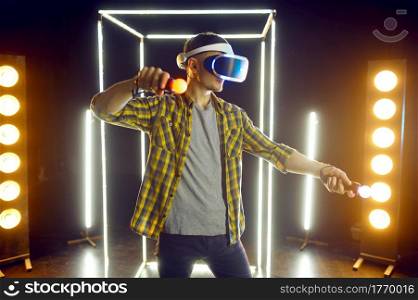 Man gaming in virtual reality headset and gamepad in luminous cube. Dark playing club interior, spotlight on background, VR technology with 3D vision. Man gaming in virtual reality headset and gamepad