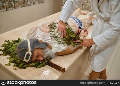 Man getting back massage with oak broom in bath. Spa, hammam or russian bathhouse therapy. Man getting massage with broom in bath