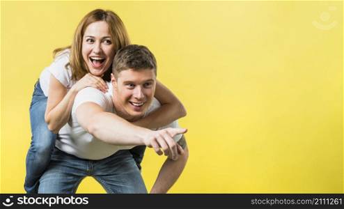 man giving her laughing girlfriend piggyback ride pointing camera against yellow background