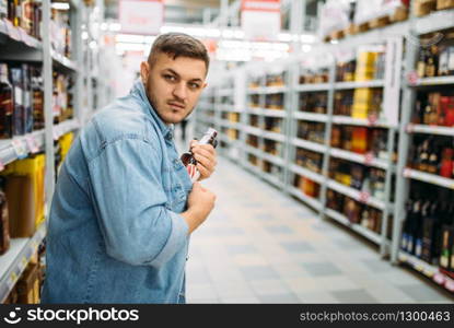 Man hides a bottle of alcohol under his shirt in supermarket. Male thief in shop, husband conducting a covert operation, shoplifling. Man hides bottle of alcohol under his shirt