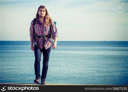 Man hiker backpacker walking with backpack by seaside at sunny day. Adventure, summer, tourism active lifestyle. Young long haired guy tramping