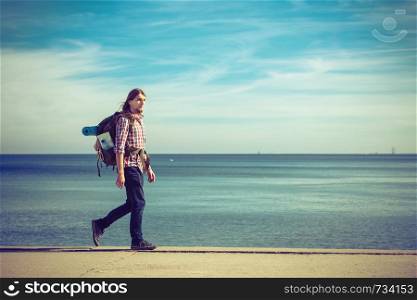 Man hiker backpacker walking with backpack by seaside at sunny day. Adventure, summer, tourism active lifestyle. Young long haired guy tramping. Man hiker with backpack tramping by seaside