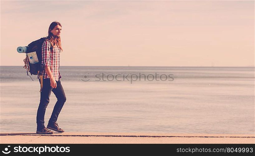 Man hiker backpacker walking with backpack by seaside at sunny day. Adventure, summer, tourism active lifestyle. Young long haired guy tramping