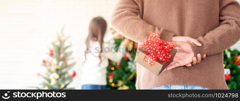 Man hold christmas gift behind his back for surprise his girlfriend while she decorating christmas tree in Chgristmas holiday season greeting. Panoramic crop using as web banner