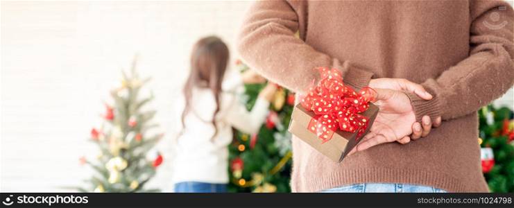 Man hold christmas gift behind his back for surprise his girlfriend while she decorating christmas tree in Chgristmas holiday season greeting. Panoramic crop using as web banner