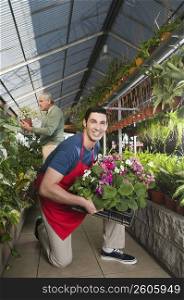 Man holding a crate of plants in a garden center with a customer in the background