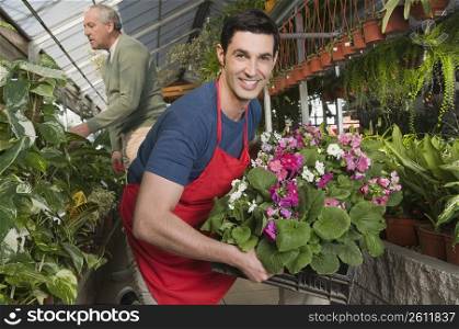 Man holding a crate of plants in a garden center with a customer in the background