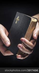 Man holding an old small black bible in his hands. Short depth of field, the sharpness is in the cross.