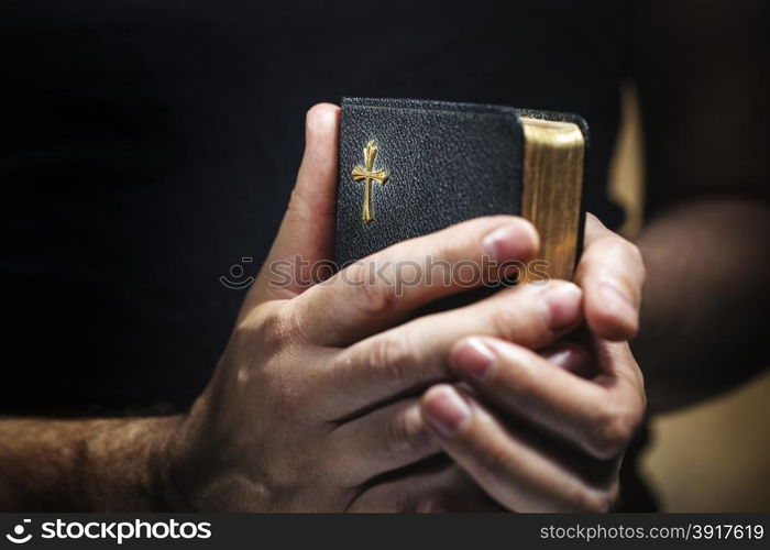 Man holding an old small black bible in his hands. Short depth of field, the sharpness is in the cross.