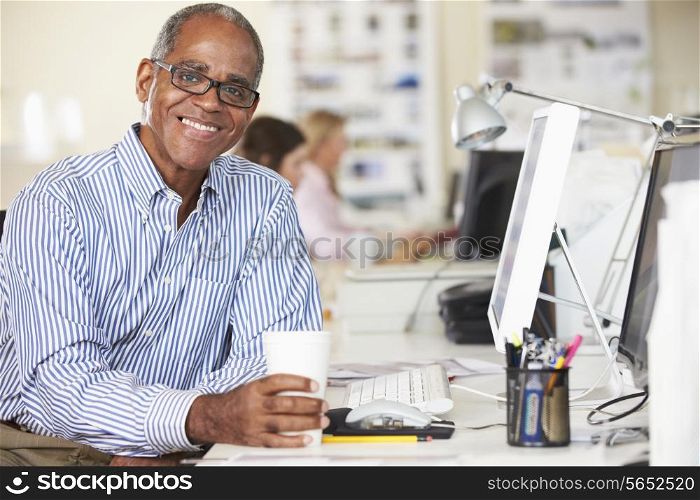 Man Holding Cup Working At Desk In Busy Creative Office