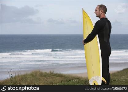 Man holding surfboard on hill looking at sea, side view
