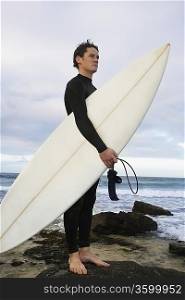 Man holding surfboard on rocks on beach, side view