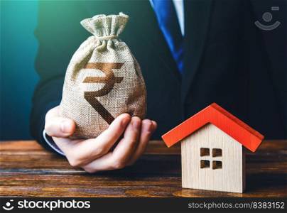 Man holds out a indian rupee bag near the house. Property appraisal. Favorable terms and conditions, low interest rate. Bank approval for issuing a mortgage loan. Home purchase, invest in real estate.