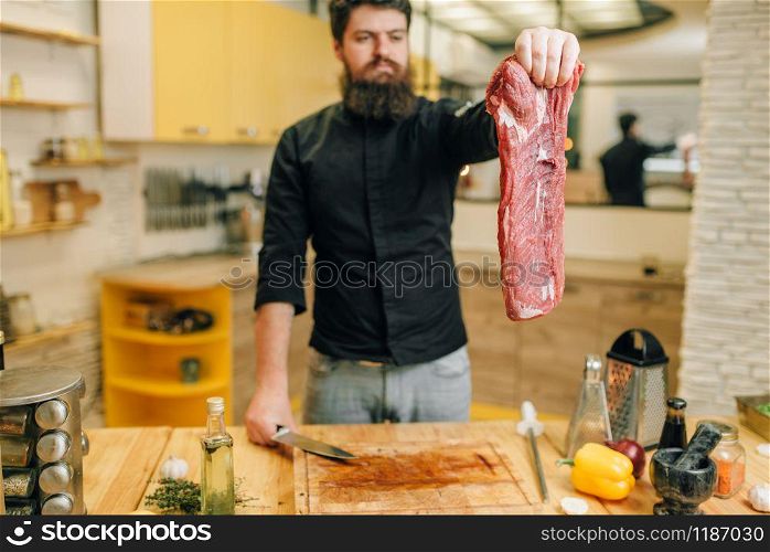 Man holds piece of fresh raw meat, kitchen interior on background. Chef cooking tenderloin with vegetables, spices and herbs