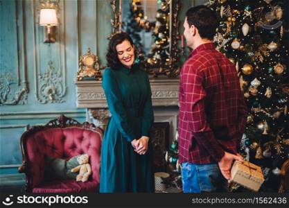 Man holds present behind back going to give it to his wife, who is standing near decorated beautiful New Year tree. Pleased shy woman going to recieve gift from husband, being glad and excited
