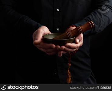 man in a black shirt holds a Tibetan brass singing bowl and a wooden stick, a ritual of meditation, prayers and immersion in a trance. Alternative treatment