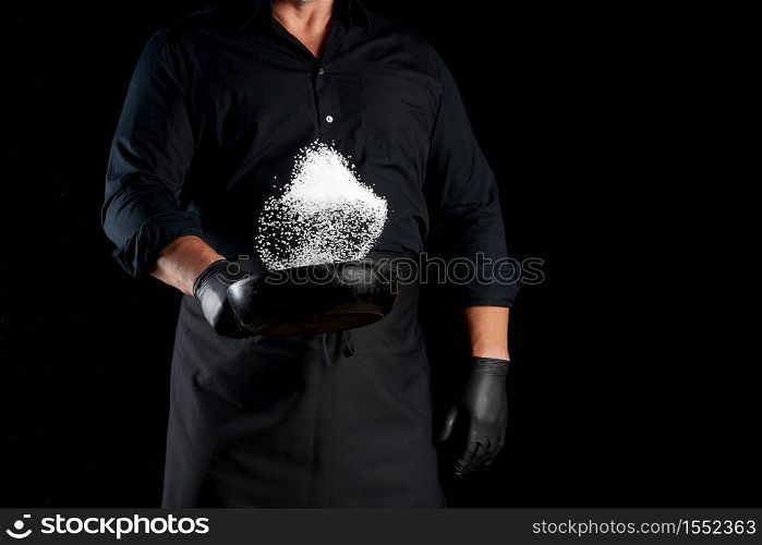 man in a black uniform holding a round cast iron pan with salt, the chef tosses white salt up on a black background