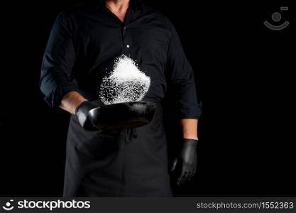 man in a black uniform holding a round cast iron pan with salt, the chef tosses white salt up on a black background