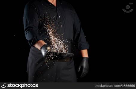 man in a black uniform holding a round cast iron pan with salt and pepper, chef tosses spice up on a black background, copy space