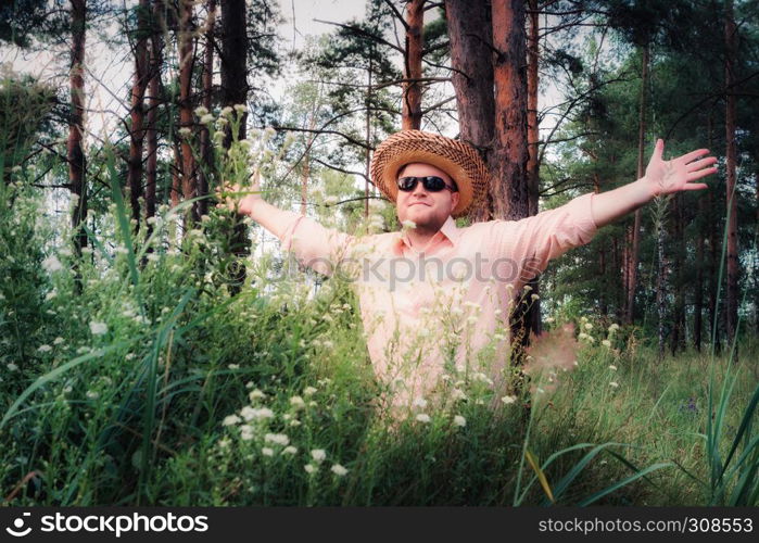 Man in a straw hat with arms raised sitting among flowers on the forest lawn at sunny day. Selective focus, vintage film filter.. Man Raised Arms on the Forest Lawn