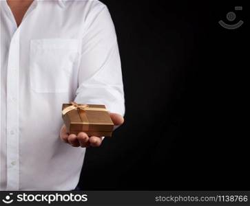 man in a white shirt holds in his left hand a closed little golden box on a dark background, concept proposal of marriage, congratulations on the holiday, copy space