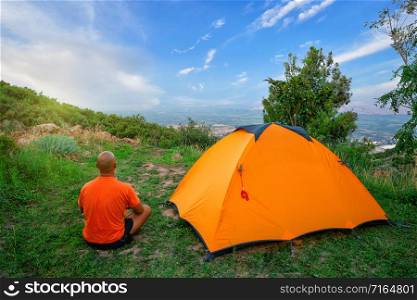Man in an orange T-shirt sits near a tent on a hill. Blue sky, bright sun.. Man in an orange T-shirt sits near tent on hill