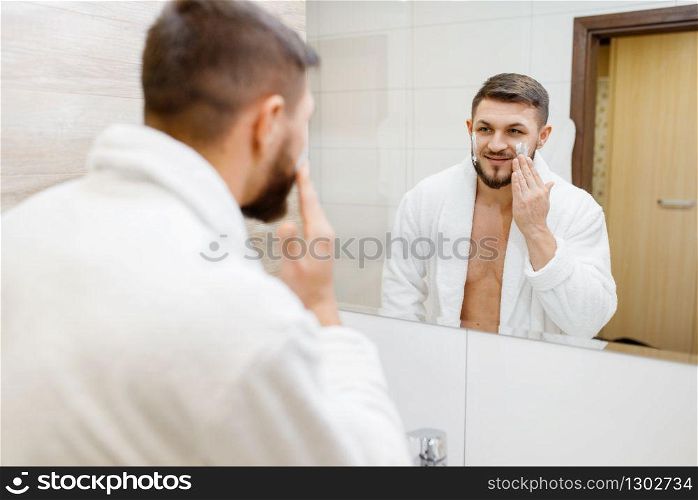 Man in bathrobe applies shaving foam at the mirror in bathroom, routine morning hygiene. Male person at the sink performs skin and body treatment procedures. Man applies shaving foam, morning hygiene