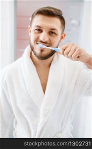 Man in bathrobe brushes his teeth in bathroom, routine morning hygiene. Male person at the sink performs skin and body treatment procedures. Man in bathrobe brushes his teeth in bathroom