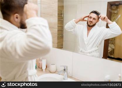Man in bathrobe combs his hair in bathroom, routine morning hygiene. Male person at the sink performs skin and body treatment procedures. Man in bathrobe combs his hair in bathroom