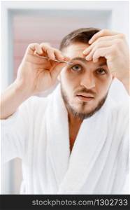 Man in bathrobe removes eyebrow hair in bathroom, routine morning hygiene. Male person at the sink performs skin and body treatment procedures. Man removes eyebrow hair, morning hygiene