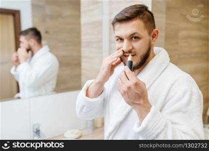 Man in bathrobe removes nose hair in bathroom, routine morning hygiene. Male person at the sink performs skin and body treatment procedures. Man removes nose hair in bathroom, morning hygiene