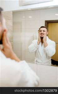 Man in bathrobe rubs aftershave over his face at the mirror in bathroom, routine morning hygiene. Male person at the sink performs skin and body treatment procedures. Man rubs aftershave over his face, morning hygiene