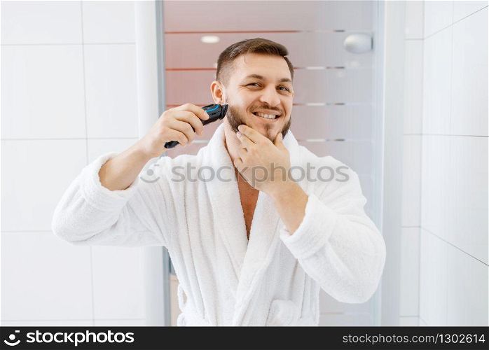 Man in bathrobe shaves his beard with an electric razor in bathroom, routine morning hygiene. Male person at the sink performs skin and body treatment procedures. Man shaves his beard with an electric razor