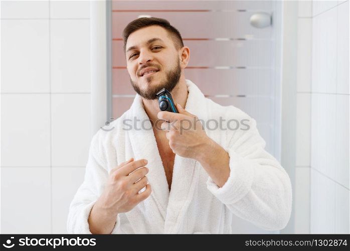 Man in bathrobe shaves his beard with an electric razor in bathroom, routine morning hygiene.. Man shaves his beard with an electric razor