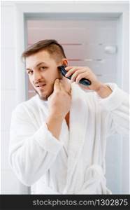 Man in bathrobe shaves his beard with an electric razor in bathroom, routine morning hygiene.. Man shaves his beard with an electric razor