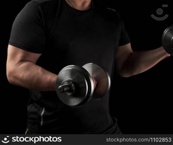 man in black clothes holds steel dumbbells in his hands, his muscles are tense, low key, sports background