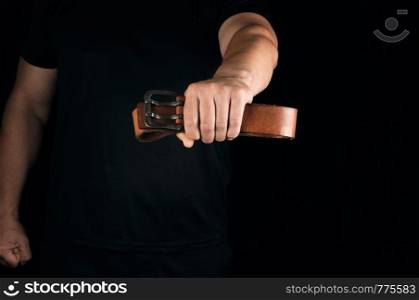man in black clothes is holding a brown leather belt with a buckle, concept of aggression and domestic violence