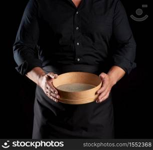 man in black uniform holding empty vintage round wooden sieve for sifting flour, chef stands against black background