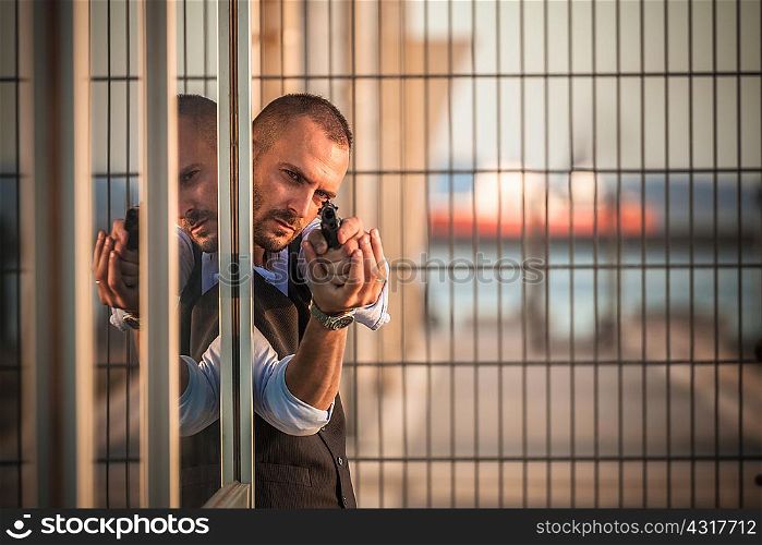 Man in business attire poised behind corner in harbour pointing handgun, Cagliari, Sardinia, Italy