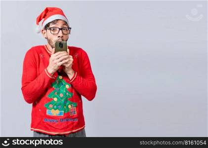Man in christmas hat with amazed face holding phone. Online Christmas special offers concept. Young man in christmas clothes and hat amazed holding the phone
