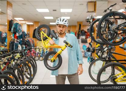 Man in cycling helmet holds children&rsquo;s bicycle, shopping in sports shop. Summer season extreme lifestyle, active leisure store, customer buying cycle and equipment for family riding