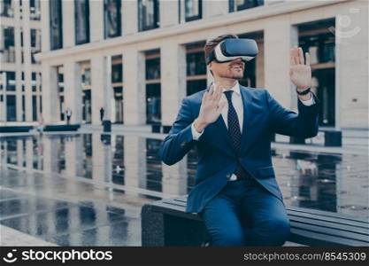 Man in formal outfit in VR goggles, trying to reach something in virtual reality with his hands in front of him, sitting on bench in front of fountain and office buildings outdoors during coffee break. Man in VR goggles trying to reach something in virtual reality with his hands while sitting outdoors