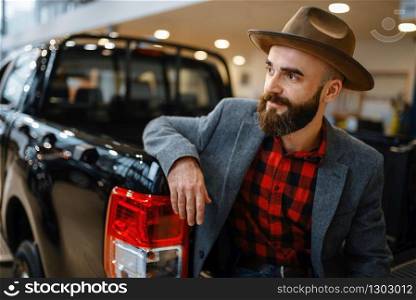 Man in hat poses at new pickup truck in car dealership. Customer in vehicle showroom, male person buying transport, auto dealer business. Man in hat poses at pickup truck in car dealership